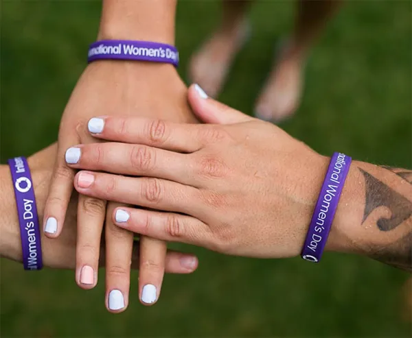 Three hands stacked together wearing purple International Women's Day bracelets