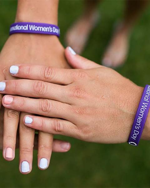 Three hands stacked together wearing purple International Women's Day bracelets